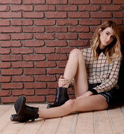 A woman with long hair sits on a wooden floor against a red brick wall, wearing a grid-patterned blouse, black shorts, and the Isla sleek black heeled booties crafted from smooth leather by Bed|Stü.