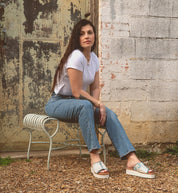 A woman wearing the Fairlee II flatform leather slide sandals by Bed Stu sits on a metal bench outdoors in a white t-shirt and jeans, with a distressed wall and door in the background.