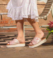 A person stands on a checkered floor near a metal chair and pink flower, wearing Bed Stu Fairlee II pink platform slide sandals with cushioned footbed and a white dress.