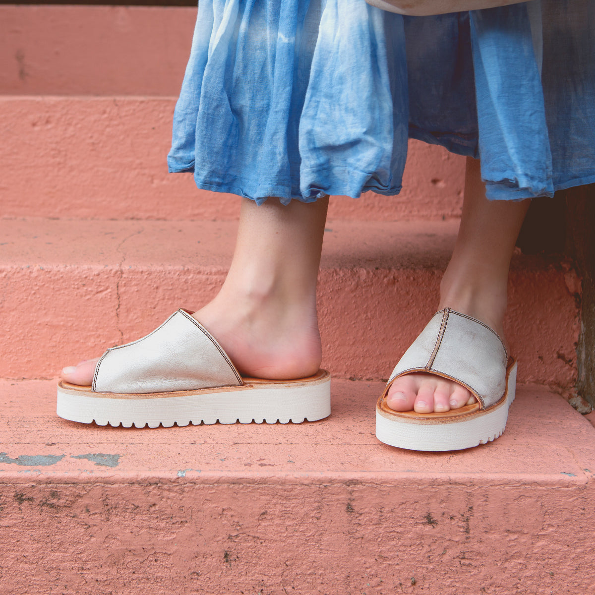 A person wearing Bed|Stü Fairlee II white slip-on platform slide sandals with a cushioned footbed and a blue tie-dye skirt stands on pink concrete steps.