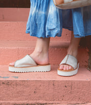 A person wearing Bed|Stü Fairlee II white slip-on platform slide sandals with a cushioned footbed and a blue tie-dye skirt stands on pink concrete steps.