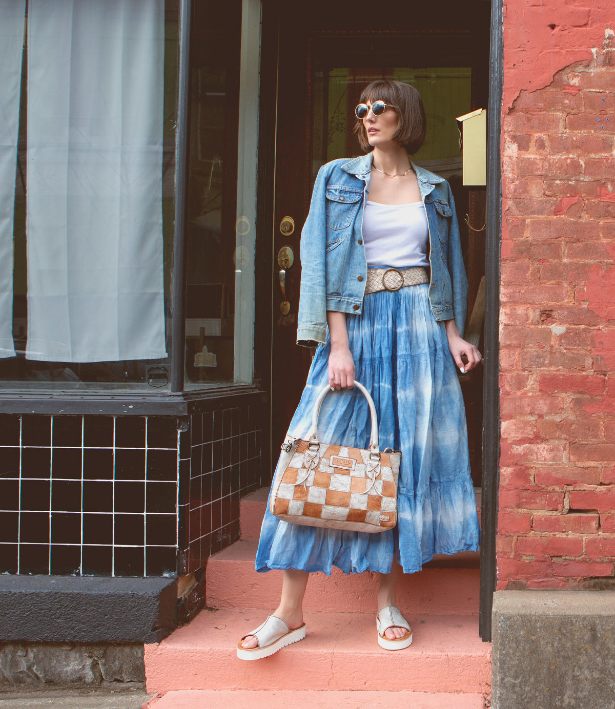 A woman in a denim jacket, white top, and blue tie-dye skirt stands on pink steps near a brick wall and tiled storefront, holding a checkered handbag and wearing sunglasses along with stylish Bed|Stü Fairlee II leather flatform sandals featuring a cushioned footbed.