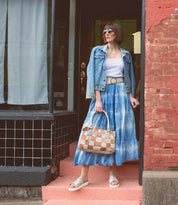 A woman in a denim jacket, white top, and blue tie-dye skirt stands on pink steps near a brick wall and tiled storefront, holding a checkered handbag and wearing sunglasses along with stylish Bed|Stü Fairlee II leather flatform sandals featuring a cushioned footbed.