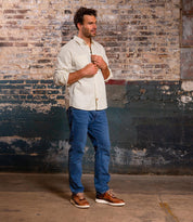 A man stands indoors near a weathered brick wall, wearing blue jeans, a light beige shirt, and Bed|Stü Fairman brown shoes with a lightweight sole; he looks to the side with his hands near his shirt collar.