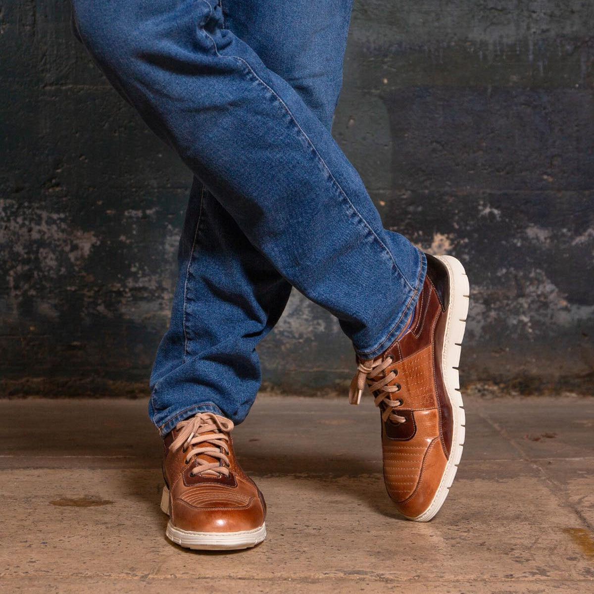A person wearing blue jeans and the Bed|Stü Fairman stylish men's sneakers in three-tone leather stands on a concrete floor with legs crossed in front of a textured dark wall.