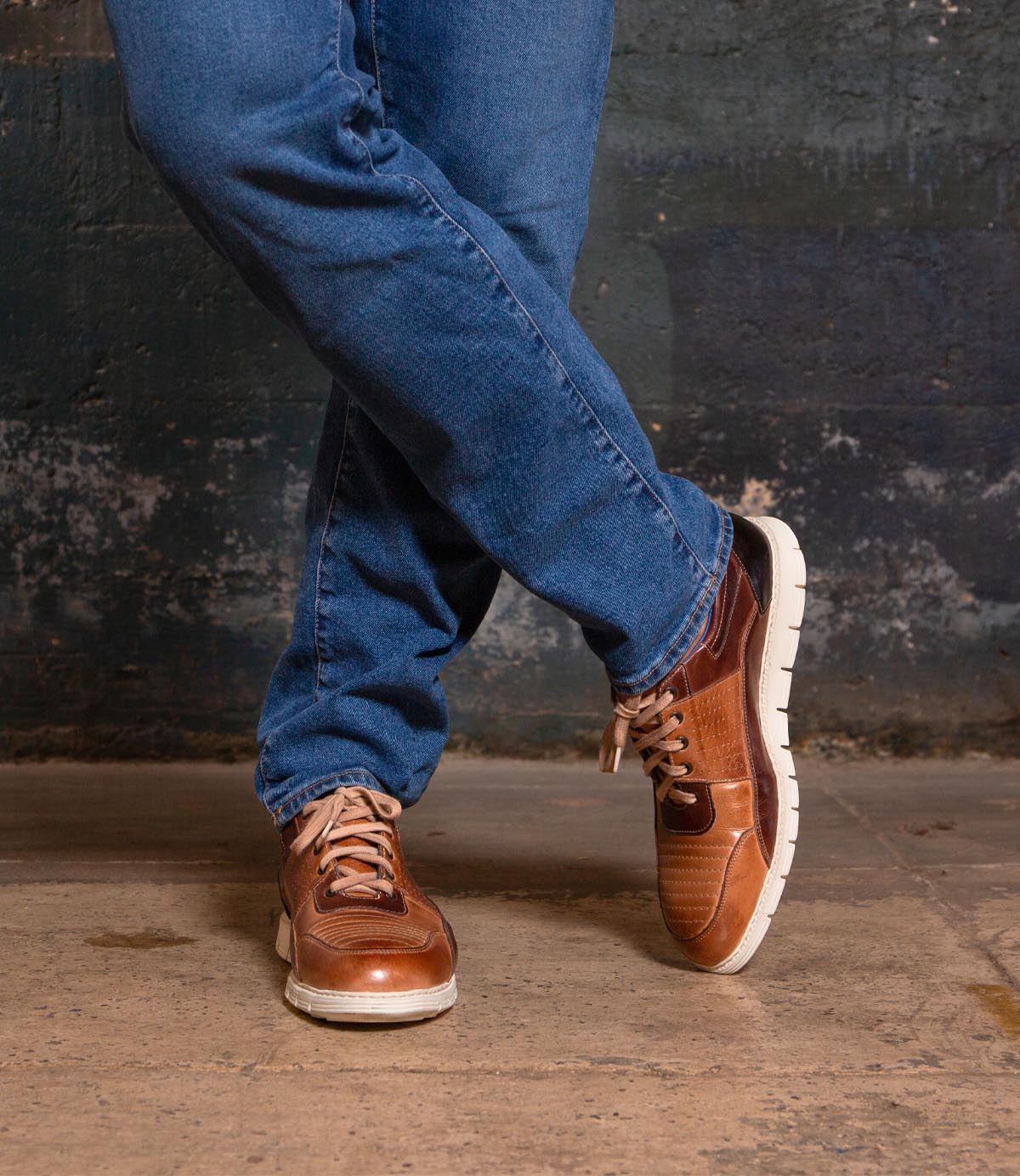 A person wearing blue jeans and the Bed|Stü Fairman stylish men's sneakers in three-tone leather stands on a concrete floor with legs crossed in front of a textured dark wall.