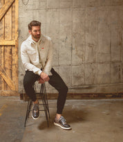 A man in a light jacket and dark pants sits on a tall stool against a concrete wall with wooden paneling to the left, looking off to the side, his look completed by the Fairman men's sneakers from Bed|Stü, crafted from three-tone leather and set on a lightweight sole.
