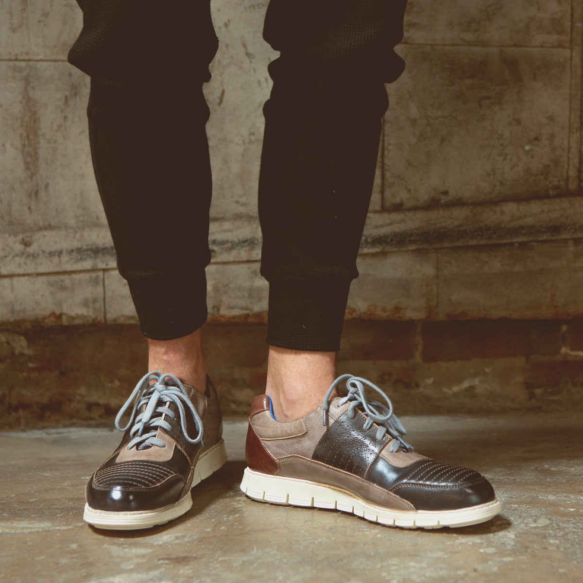 A person wearing black jogger pants and the Fairman brown men’s sneakers by Bed|Stü, featuring a lightweight sole and white accents, standing on a concrete floor against a stone wall.