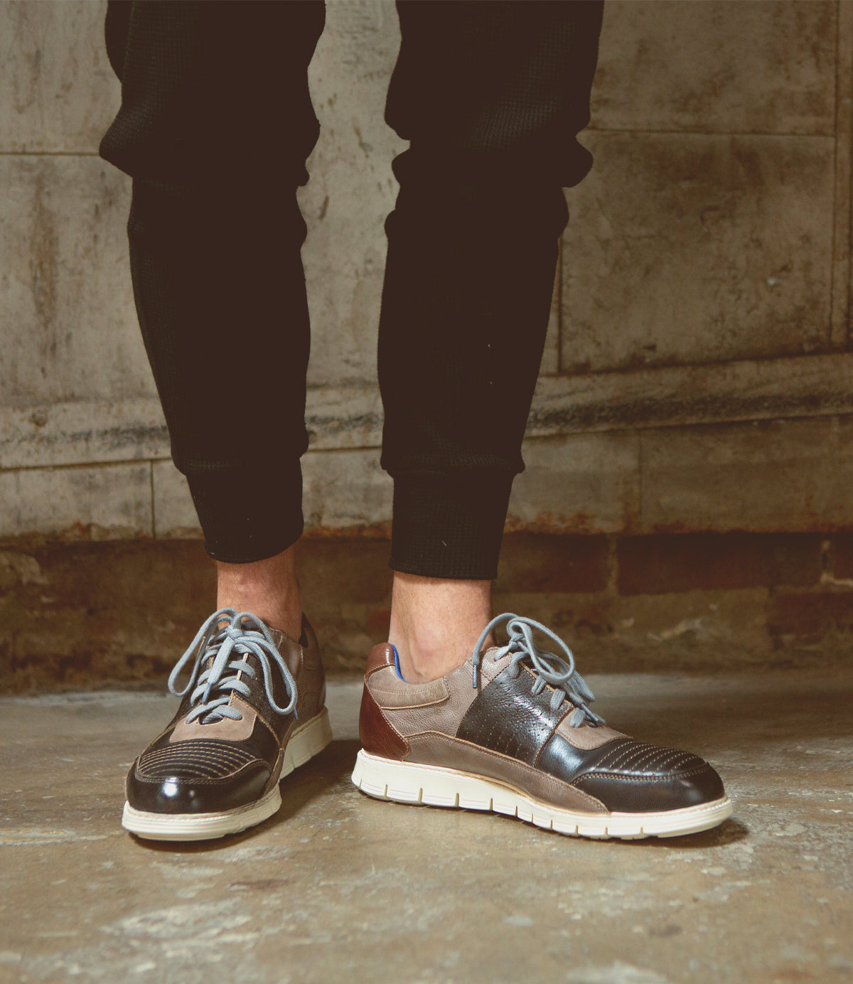 A person wearing black jogger pants and the Fairman brown men’s sneakers by Bed|Stü, featuring a lightweight sole and white accents, standing on a concrete floor against a stone wall.