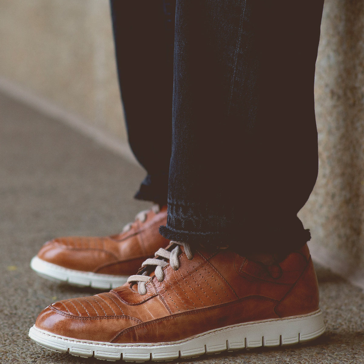 A person wearing dark jeans and the Bed|Stü Fairman men's sneaker, featuring three-tone leather and a lightweight sole, stands on a speckled surface near a stone wall.