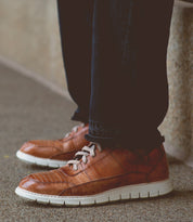 A person wearing dark jeans and the Bed|Stü Fairman men's sneaker, featuring three-tone leather and a lightweight sole, stands on a speckled surface near a stone wall.