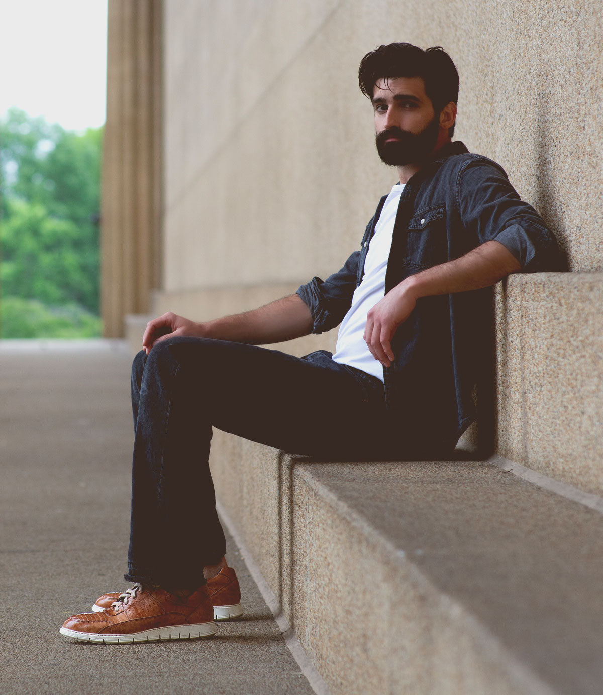 A man with dark hair and a beard sits on a stone ledge outdoors, wearing a white shirt, dark jeans, a dark jacket, and stylish Bed|Stü Fairman three-tone leather men’s sneakers with a lightweight sole. Trees and a building wall are visible in the background.