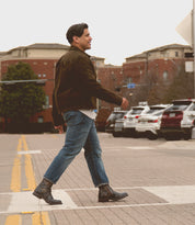 A man wearing a brown jacket, blue jeans, and Bed|Stü's Fellow rugged staple moto boots with Goodyear welt construction walks across a crosswalk on an urban street lined with parked cars and buildings.