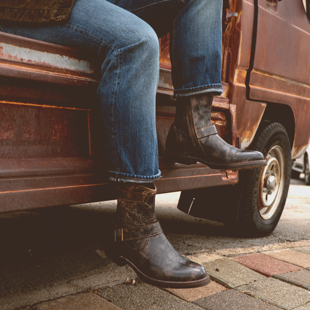 Wearing the Bed|Stü Fellow boots—crafted with durable Goodyear welt construction—a person in blue jeans sits atop the step of a rusty vehicle parked along a brick sidewalk, highlighting this rugged staple that's perfect for any adventurous outing.