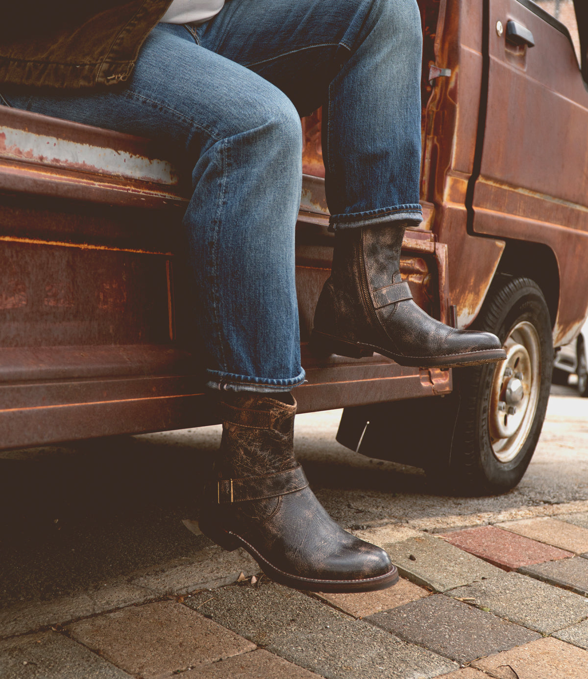 Wearing the Bed|Stü Fellow boots—crafted with durable Goodyear welt construction—a person in blue jeans sits atop the step of a rusty vehicle parked along a brick sidewalk, highlighting this rugged staple that's perfect for any adventurous outing.