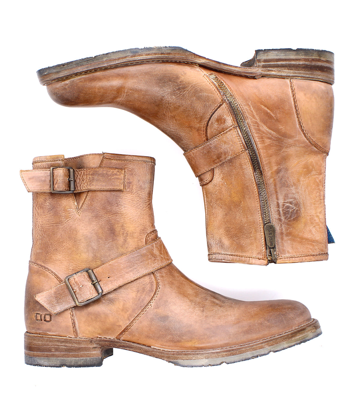 Bed|Stü's Fellow is a pair of brown leather ankle boots featuring Goodyear welt construction, side zippers, and buckle straps—a rugged staple inspired by classic moto boot design. One boot lies flat while the other rests on its side.