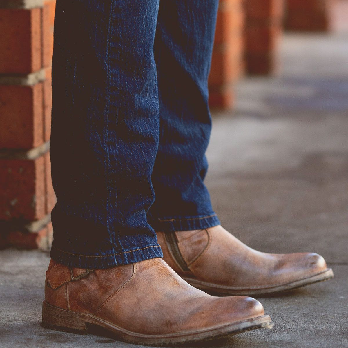 A person wearing blue jeans and rugged Bed|Stü Fellow brown leather moto boots with Goodyear welt construction stands on a concrete sidewalk near a brick wall.
