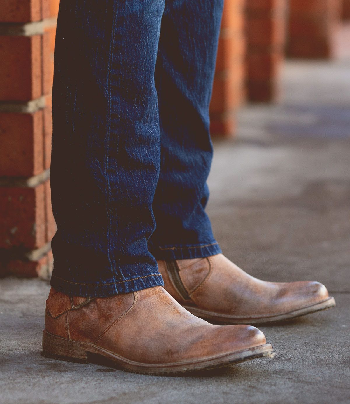 A person wearing blue jeans and rugged Bed|Stü Fellow brown leather moto boots with Goodyear welt construction stands on a concrete sidewalk near a brick wall.