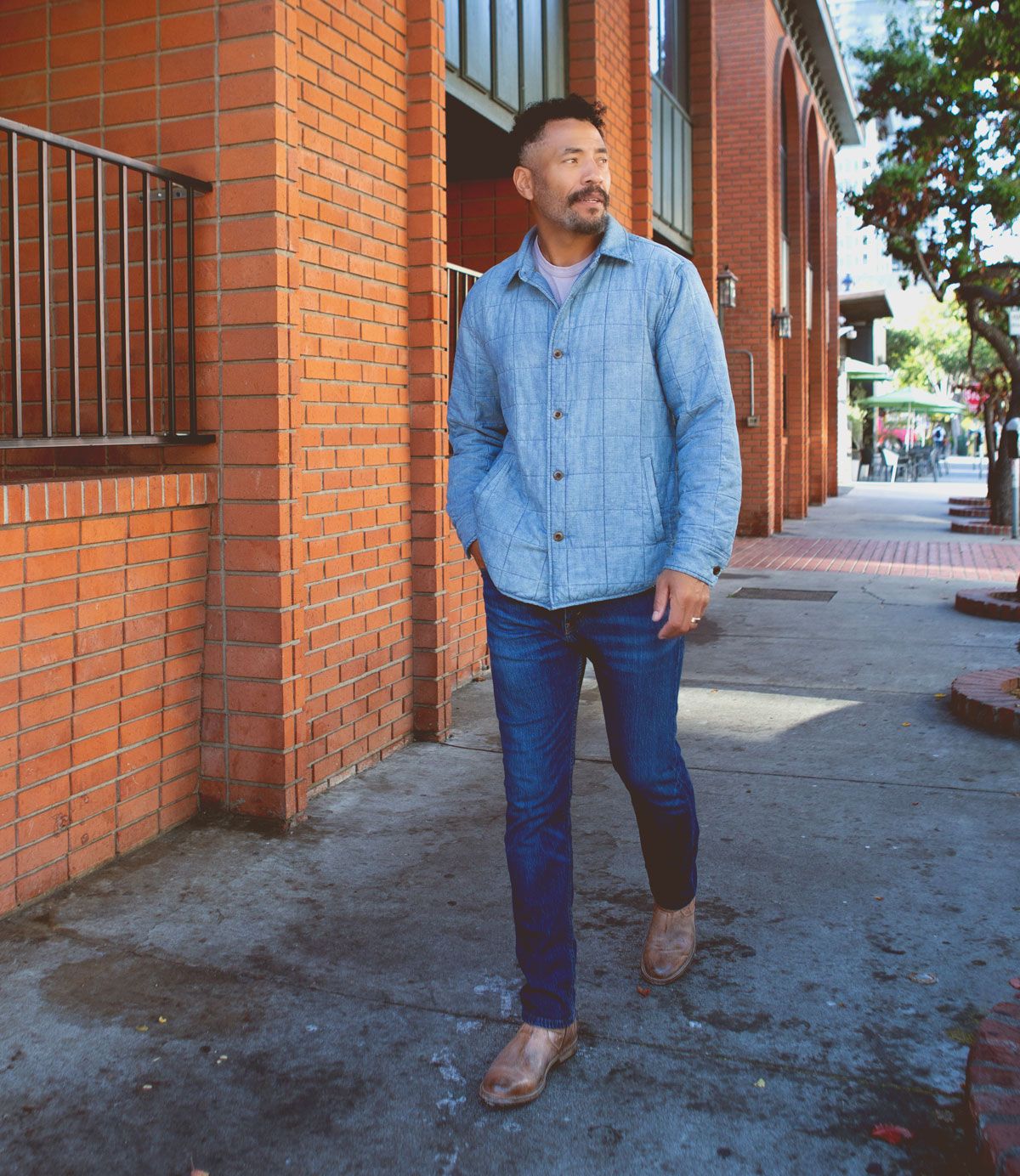 A man in a light blue button-up shirt, blue jeans, and Bed|Stü Fellow rugged brown moto boots with Goodyear welt construction walks on a city sidewalk beside a red brick building.