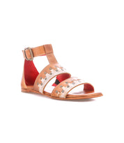 The Bed|Stü Fibril sandal features brown leather in a bohemian style, with an adjustable ankle strap, silver studs, open toe design, buckle closure, and red inner lining, photographed on a white background.