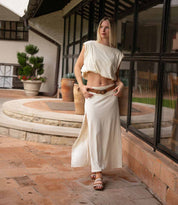A woman wearing the Fibril sandals by Bed|Stü, featuring an adjustable strap, stands on a tiled patio near large windows and potted plants. She is dressed in a cream two-piece outfit with a slit skirt.