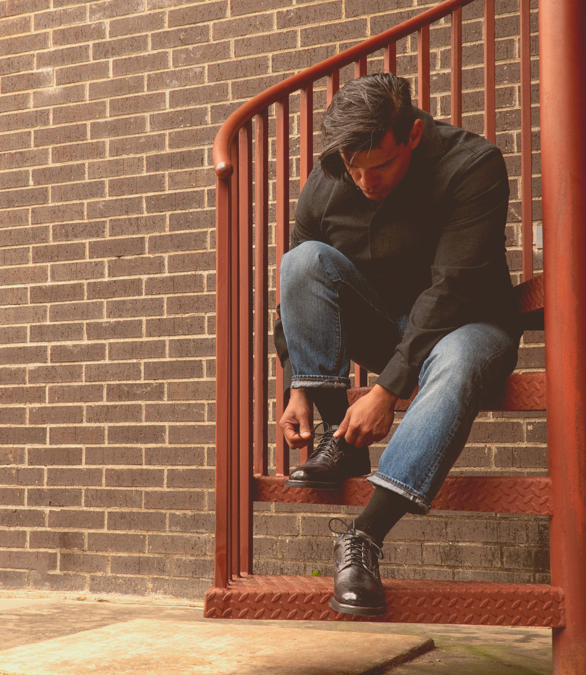 A person in jeans and a dark shirt sits on an outdoor metal stairway, bending down to tie their black Bed|Stü Fierce shoes against a brick wall backdrop.