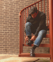 A person in jeans and a dark shirt sits on an outdoor metal stairway, bending down to tie their black Bed|Stü Fierce shoes against a brick wall backdrop.