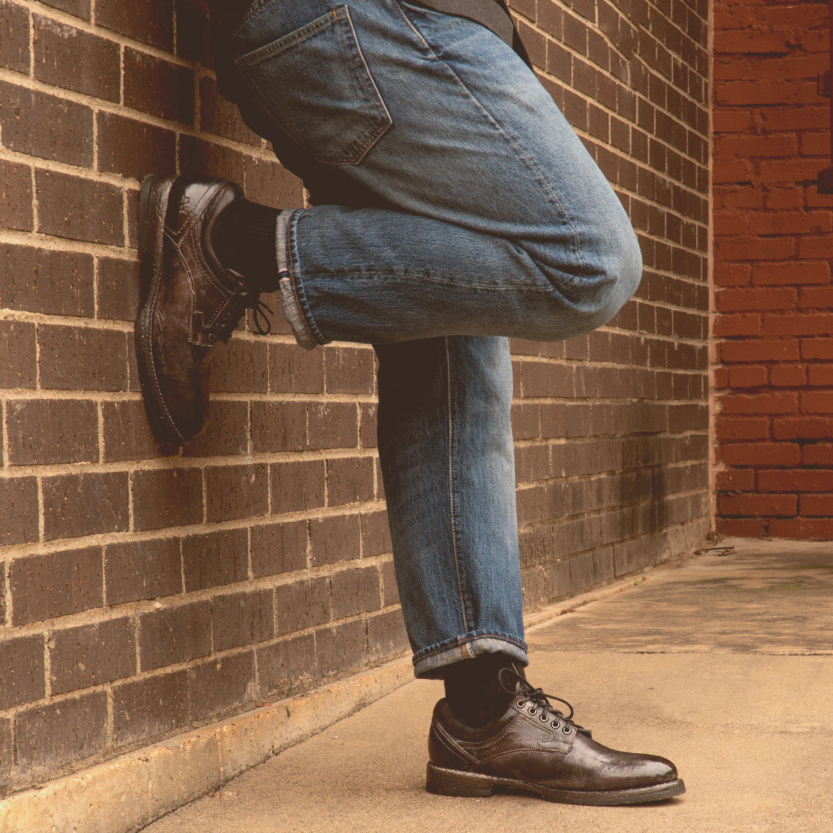Wearing Bed|Stü Fierce jeans and brown oxford shoes with a Goodyear welt, the person stands on a sidewalk with one leg crossed, leaning casually against a brick wall.