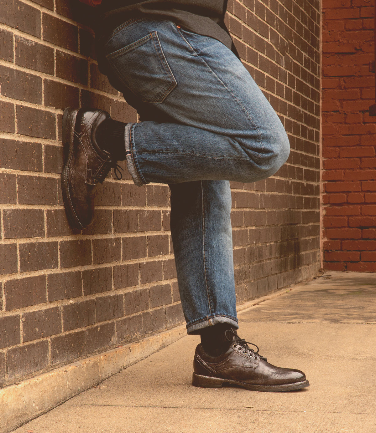 Wearing Bed|Stü Fierce jeans and brown oxford shoes with a Goodyear welt, the person stands on a sidewalk with one leg crossed, leaning casually against a brick wall.