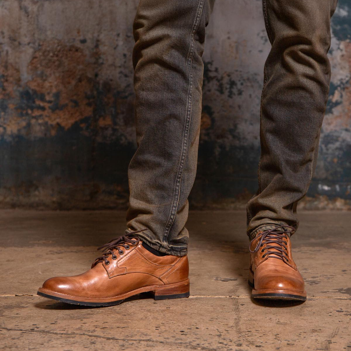 A person wearing Bed|Stü Fierce washed leather derby dress shoes with a Goodyear welt and dark jeans stands on a worn concrete floor against a distressed wall.