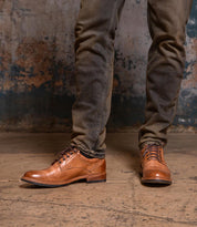 A person wearing Bed|Stü Fierce washed leather derby dress shoes with a Goodyear welt and dark jeans stands on a worn concrete floor against a distressed wall.
