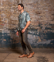 A man stands indoors near a worn brick wall, wearing a patterned green shirt, dark jeans, and Bed|Stü Fierce brown dress shoes featuring a jute-infused outsole.