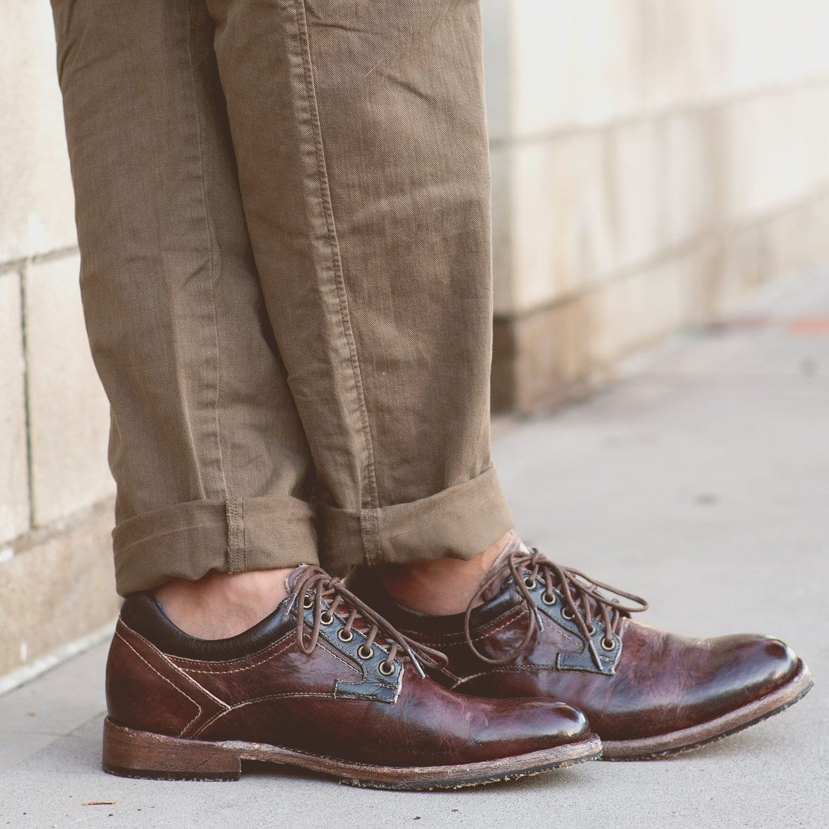 A person wearing Bed|Stü Fierce brown washed leather derby dress shoes with a Goodyear welt and light brown pants stands on a sidewalk next to a brick wall.