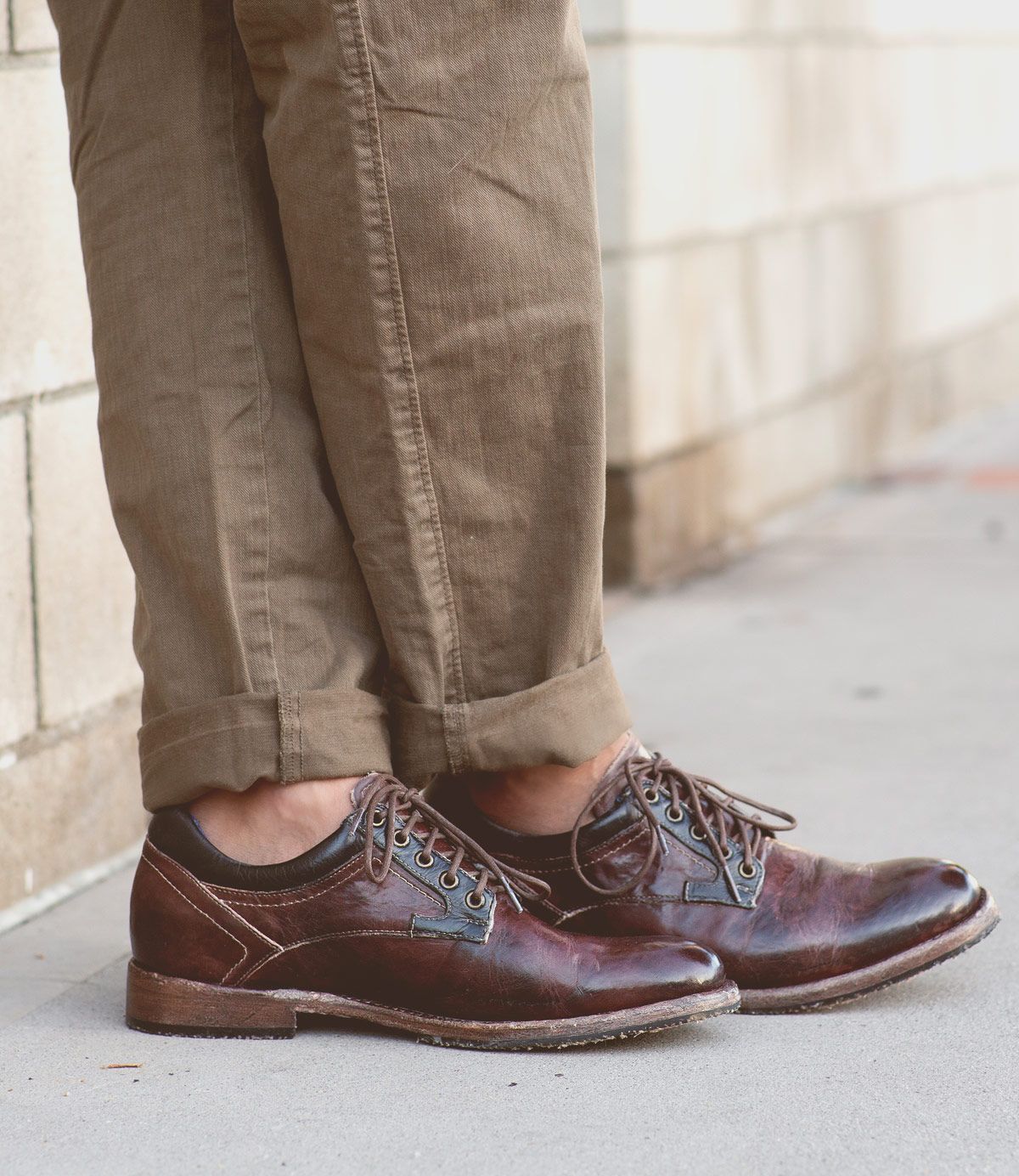 A person wearing Bed|Stü Fierce brown washed leather derby dress shoes with a Goodyear welt and light brown pants stands on a sidewalk next to a brick wall.