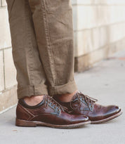 A person wearing Bed|Stü Fierce brown washed leather derby dress shoes with a Goodyear welt and light brown pants stands on a sidewalk next to a brick wall.