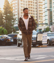 A man walks through a parking lot, sporting a patterned jacket, white shirt, and brown pants, paired with the Bed|Stü Fierce washed leather derby shoes featuring a Goodyear welt; buildings and parked cars can be seen in the background.