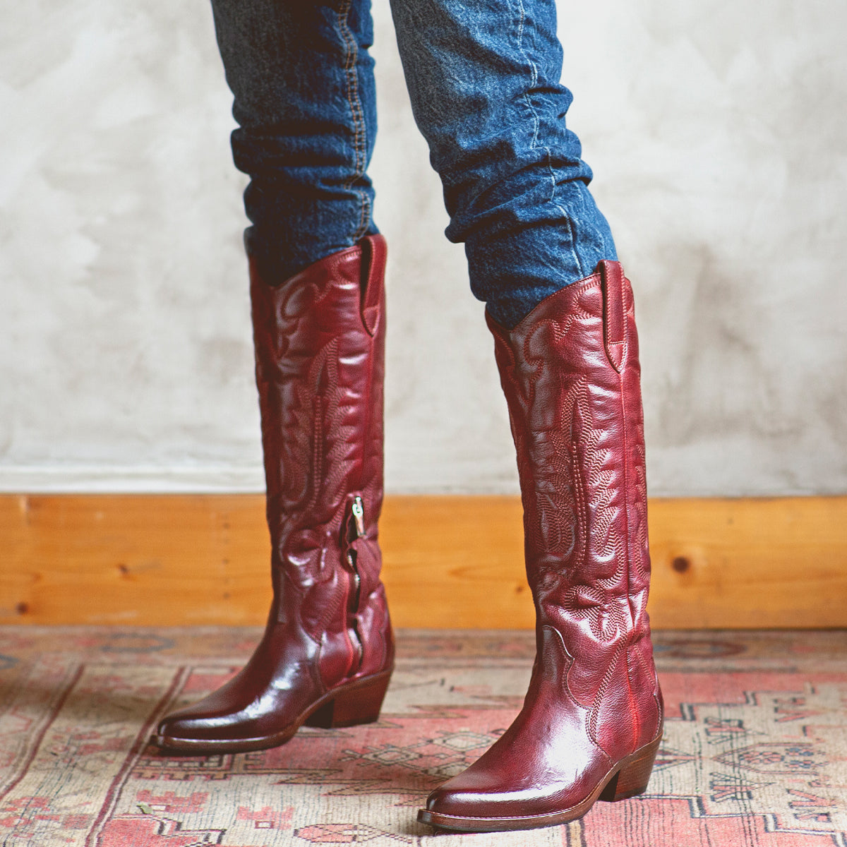 Wearing Bed|Stü's Finito tall, dark red leather cowboy boots with a classic western style, a person in blue jeans stands on a patterned rug against a light-colored wall.