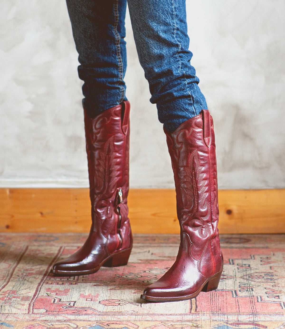 Wearing Bed|Stü's Finito tall, dark red leather cowboy boots with a classic western style, a person in blue jeans stands on a patterned rug against a light-colored wall.