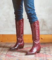 Wearing Bed|Stü's Finito tall, dark red leather cowboy boots with a classic western style, a person in blue jeans stands on a patterned rug against a light-colored wall.