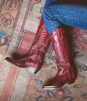 A person wearing Bed|Stü Finito red leather cowboy boots with a distressed finish and blue jeans sits cross-legged on a patterned rug.
