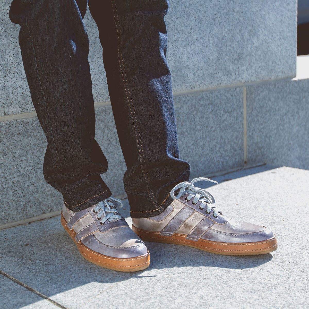 A person wearing blue jeans and Bed|Stü Firepower gray sneakers with white laces and leather stripe details stands on a light gray concrete surface near a stone wall.