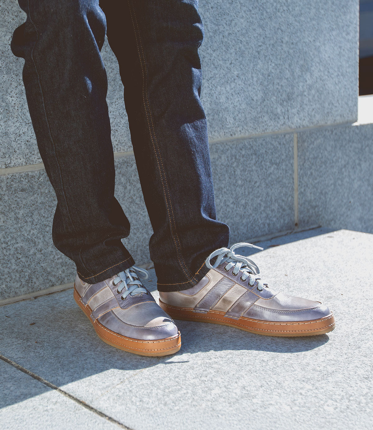 A person wearing blue jeans and Bed|Stü Firepower gray sneakers with white laces and leather stripe details stands on a light gray concrete surface near a stone wall.