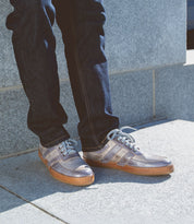 A person wearing blue jeans and Bed|Stü Firepower gray sneakers with white laces and leather stripe details stands on a light gray concrete surface near a stone wall.