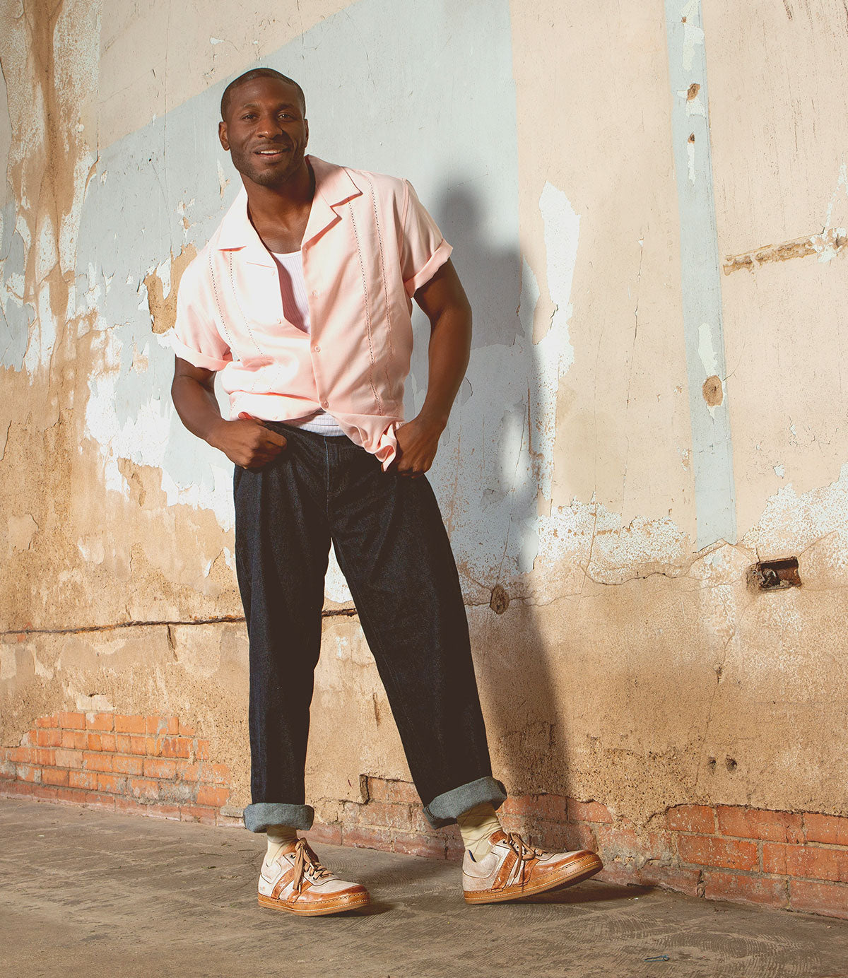 A man stands indoors against a worn, textured wall, wearing beige Bed|Stü Firepower everyday sneakers with a textured sole, paired with a light pink short-sleeve shirt and dark cuffed pants. One hand is in his pocket as he smiles slightly.