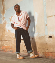 A man stands indoors against a worn, textured wall, wearing beige Bed|Stü Firepower everyday sneakers with a textured sole, paired with a light pink short-sleeve shirt and dark cuffed pants. One hand is in his pocket as he smiles slightly.