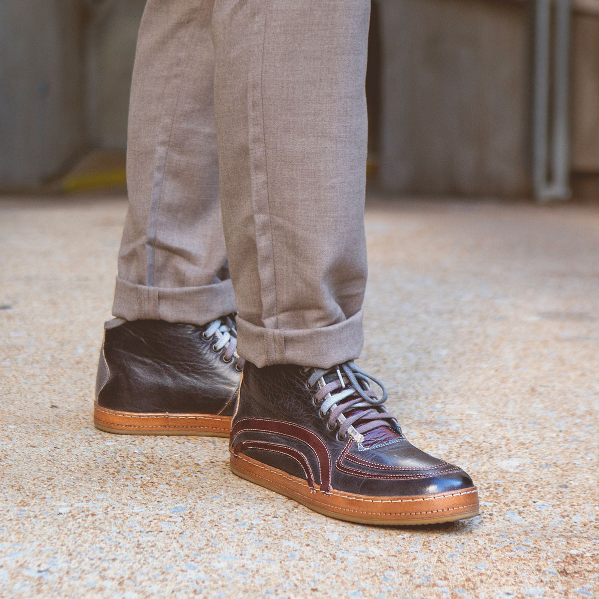 Wearing light brown pants and Bed|Stü’s Formation dark brown leather high-top sneakers—featuring detailed leather paneling inspired by vintage court styles—a person stands on textured pavement.