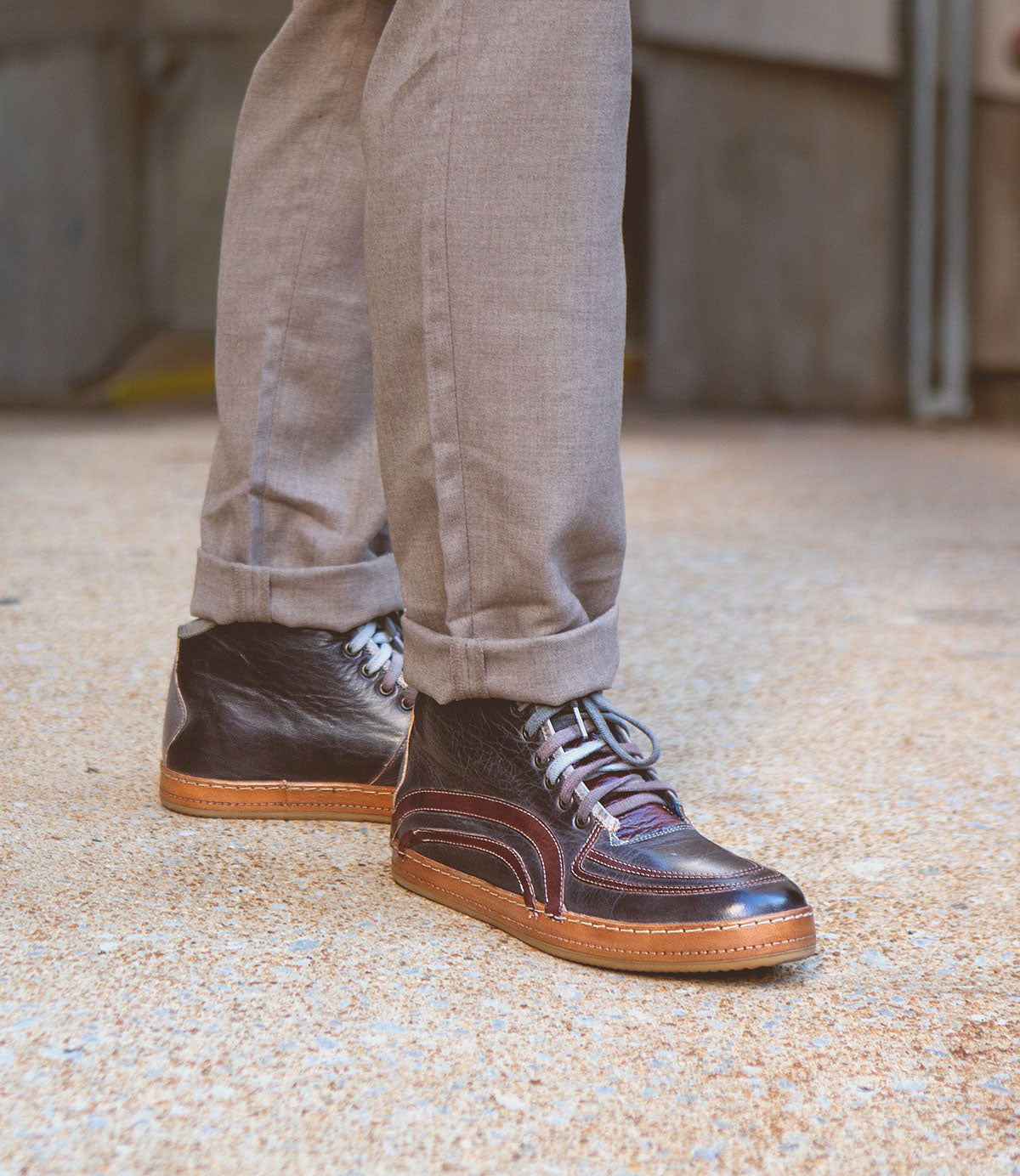 Wearing light brown pants and Bed|Stü’s Formation dark brown leather high-top sneakers—featuring detailed leather paneling inspired by vintage court styles—a person stands on textured pavement.