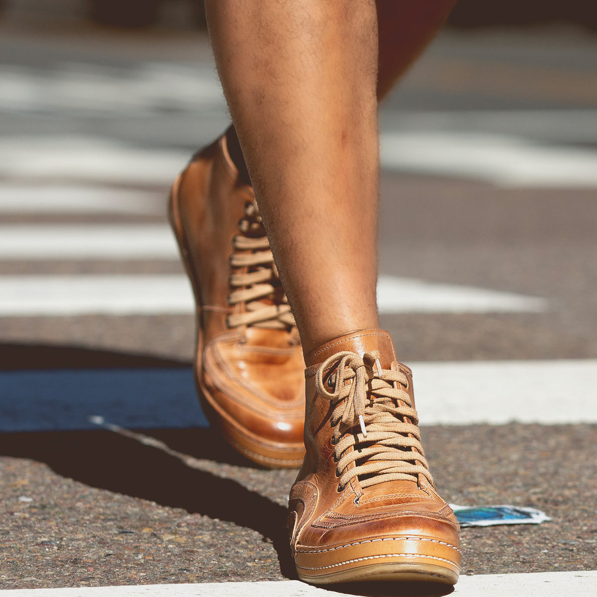 A person wearing Bed|Stü Formation tan leather high-top lace-up sneakers walks across a street crosswalk, with a blue object lying on the road nearby.