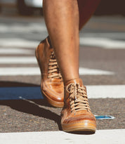 A person wearing Bed|Stü Formation tan leather high-top lace-up sneakers walks across a street crosswalk, with a blue object lying on the road nearby.