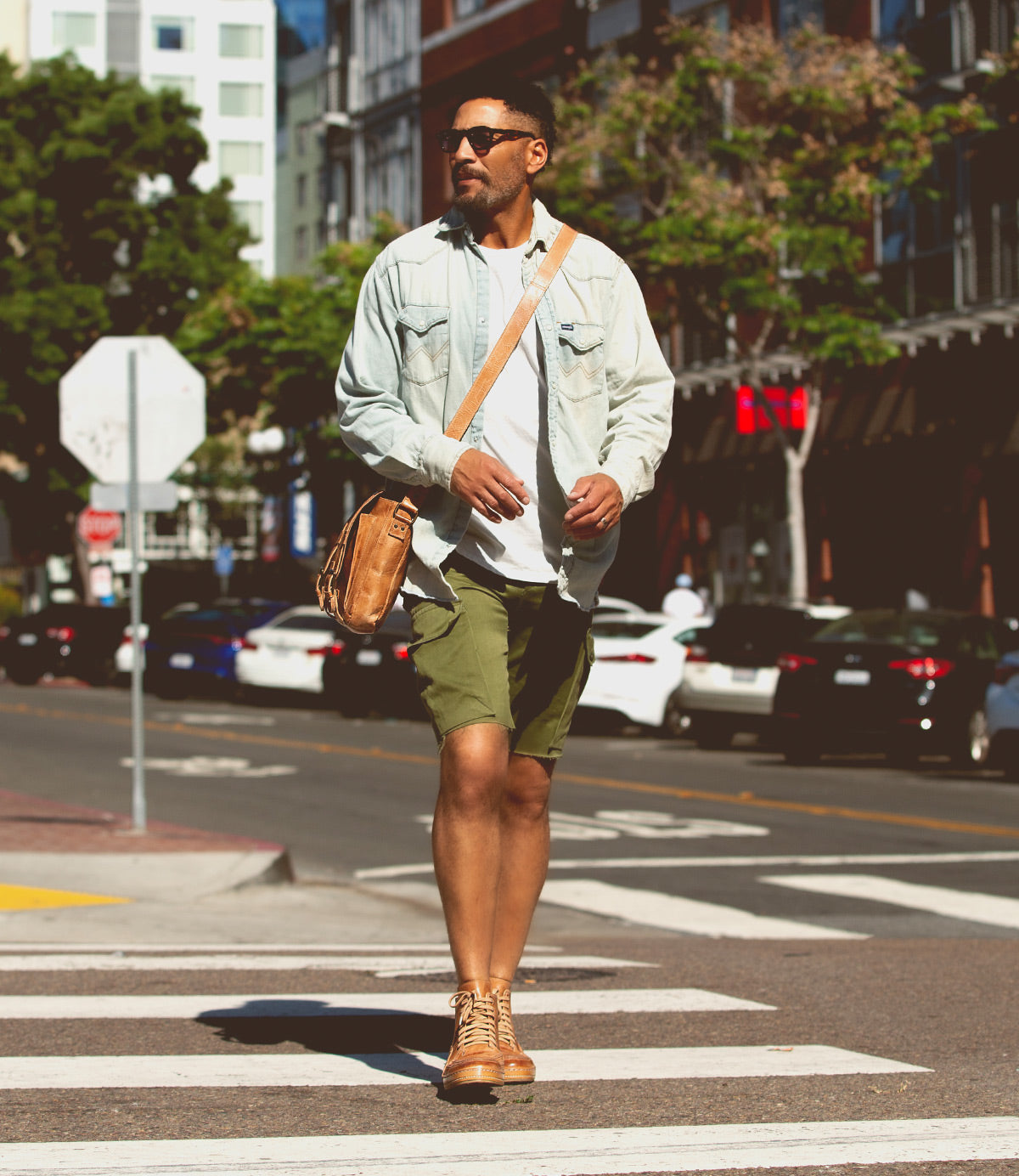 A man wearing sunglasses, a light denim shirt, green shorts, and a brown satchel walks across a city crosswalk in Bed|Stü's Formation boots—featuring distinctive leather paneling—with parked cars and buildings in the background.